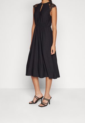 Vrouw draagt een mouwloze zwarte midijurk met kanten kapmouwtjes, een keyhole halslijn en een geplooide taille, gecombineerd met zwarte sandalen met hak.