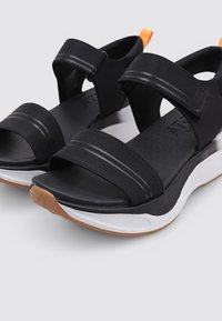 Sandalias negras con dos correas de tela ajustables, una suela acolchada y una entresuela blanca que presenta una suela de goma marrón y un acento naranja.