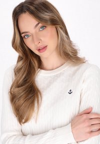 Maglione bianco a maglia rasata con colletto e polsini a costine, caratterizzato da un piccolo emblema a forma di ancora blu sul petto. Tessuto morbido e design aderente.