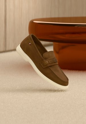 Ruskea mokkanahkainen loafer valkoisella pohjalla ja metalliyksityiskohdalla, kellumassa vaalean maton yllä kiiltävän ruskean pyöreän huonekalun vieressä.