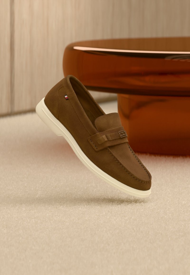 Καφέ μοκασίνι από σουέντ με λευκή σόλα και μεταλλική λεπτομέρεια, αιωρείται πάνω από ανοιχτό χαλί δίπλα σε γυαλιστερό καφέ στρογγυλό έπιπλο.