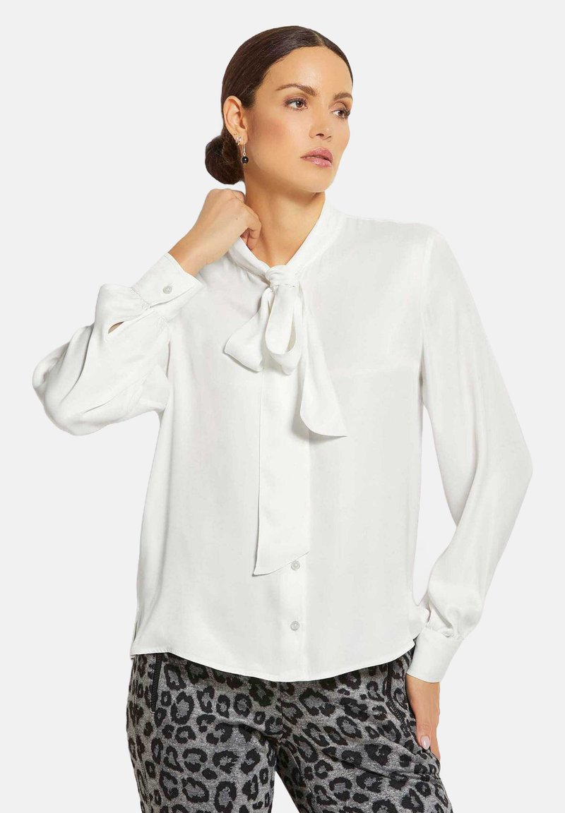 Weißes Bluse mit langen Ärmeln aus glattem Stoff, mit einer Schleife am Hals und Knopfverschlüssen an der Vorderseite. Kombiniert mit gemusterten schwarzen Hosen.