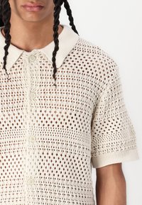 Maglietta a maniche corte lavorata a maglia in beige chiaro con motivo a trama aperta, colletto e chiusura con bottoni. Presenta dettagli testurizzati e polsini a costine.
