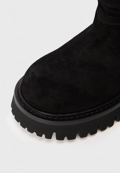 Μαύρη σουέντ μπότα με παχύ, ραχισμένο καουτσούκ πάτο και λεπτομερή ραφή στην άκρη, σε άσπρο φόντο.