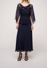 Mörkblå klänning med genomskinliga, fladdrande ärmar, spetsdetalj vid midjan och en svävande kjol. Kombineras med svarta sandaler med remmar.