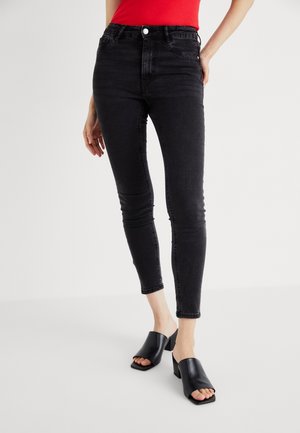 Frau trägt schwarze Skinny-Jeans, rotes ärmelloses Oberteil und schwarze offene Pumps mit Absatz, steht vor weißem Hintergrund.