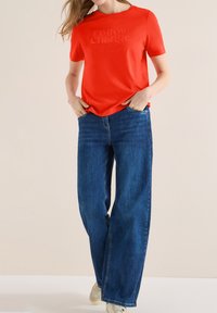 Femme portant une chemise rouge vif avec un texte, un jean bleu à jambes larges et des baskets blanches, debout avec les mains dans les poches devant un fond uni.