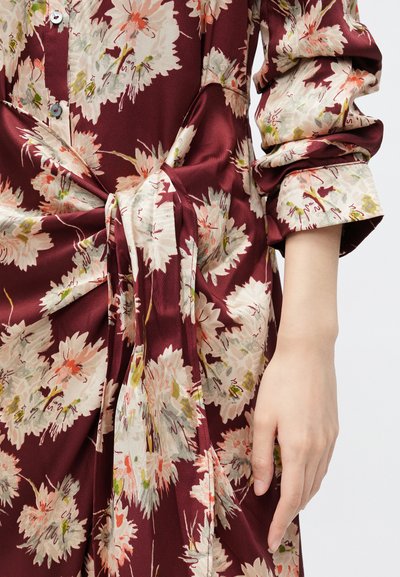 Bordowa sukienka w kwiaty wykonana z miękkiego materiału, z dużymi kremowymi kwiatami i zielonym akcentem, z wiązanym detalem w talii oraz podwiniętymi rękawami.