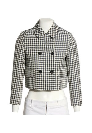Chaqueta corta de doble botonadura con patrón de pata de gallo en blanco y negro y mangas largas, mostrada en un maniquí de torso con pantalones blancos.