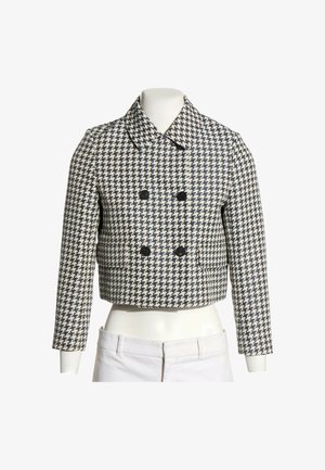 Chaqueta corta de doble botonadura con patrón de pata de gallo en blanco y negro y mangas largas, mostrada en un maniquí de torso con pantalones blancos.