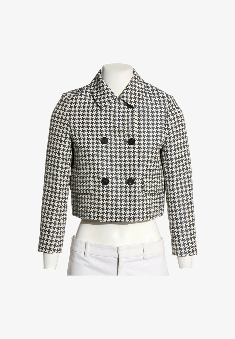 Veste courte croisée avec motif pied-de-poule noir et blanc et manches longues, présentée sur un mannequin torse avec pantalon blanc.