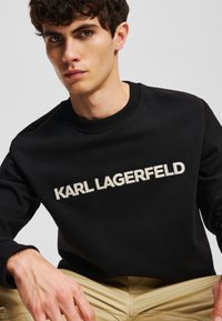 Fekete pamut pulóver, kerek nyakkivágással, bordás mandzsettaakkal és a melltartón elhelyezett, feltűnő bézs "KARL LAGERFELD" felirattal.