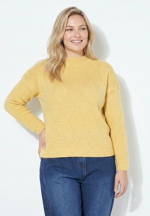 Vrouw met schouderlang blond haar, een gele gestructureerde trui en blauwe spijkerbroek, glimlachend en poserend met één hand op de heup.
