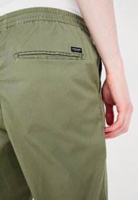 Olivgröna byxor med elastisk midja, med en bakficka med dragkedja och en liten varumärkesetikett sydd på.