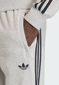 Sive trenirka hlače s crnim dizajnom s tri pruge. Imaju bočni džep i izvezeni logo. Izrađene od mekane mješavine pamuka.