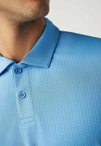 Hellblaues Poloshirt aus strukturiertem Stoff mit einem subtilen Karomuster, zwei blauen Knöpfen und einem traditionellen Kragen. 