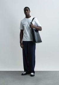 Νεαρός άνδρας που φοράει λευκό μπλουζάκι με γραφικά και σκούρο παντελόνι με ρίγες, κρατώντας μια μαύρη δερμάτινη τσάντα ώμου, στέκεται σε γκρι πάτωμα.