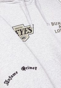 Gray hoodie with two printed designs: a central logo saying "Mud City Eyes" and text "Buried At Last" embroidered in black. Soft texture.