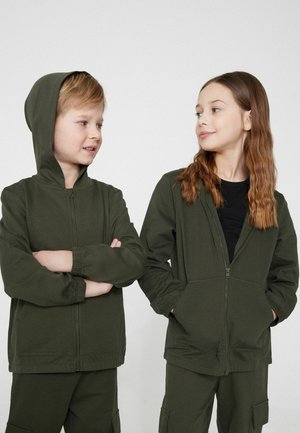 Zwei Kinder tragen passende olivgrüne Kapuzenjacken mit Reißverschluss und Hosen und sehen sich vor einem einfarbigen hellen Hintergrund an.