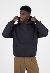 Marineblauwe hoodie van zachte stof met een voorzak, verstelbare trekkoordkap en een relaxte pasvorm. Model draagt beige broek.