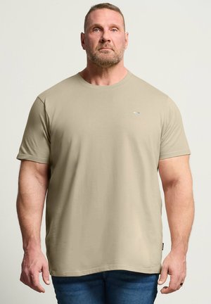 Muskularny mężczyzna z krótkimi włosami i brodą, ubrany w gładki beżowy krótki T-shirt i niebieskie dżinsy na neutralnym tle.