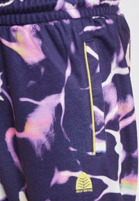 Mīksta auduma sporta bikses ar purpura pamatni, kurā ir abstrakti rozā un dzelteni raksti. Satur dzeltenu akcentu kabatas šuvē.