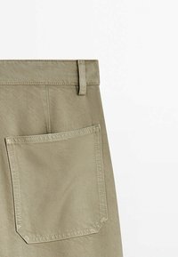 Gros plan sur la poche arrière beige d’un pantalon avec des coutures visibles et une boucle de ceinture sur un fond blanc uni.