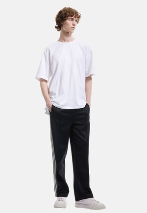 Camiseta blanca de algodón con corte holgado, combinada con pantalones de chándal negros que tienen franjas laterales blancas y zapatillas blancas, de pie contra un fondo liso.