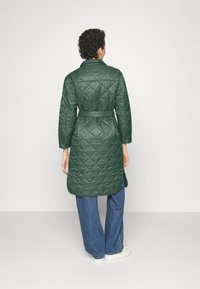 Persona vistiendo un abrigo acolchado verde oscuro con cinturón, jeans azules y zapatillas blancas, de pie frente a un fondo gris claro liso.