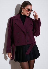 Burgundska skrajšana dvoredna jakna z velikimi črnimi gumbi, širokimi reverji in pasom; v kombinaciji s črnimi plisiranimi krili.