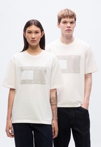 Βαμβακερά μπλουζάκια σε κρεμ απόχρωση με ανάγλυφο λογότυπο "TOMMY JEANS" σε ορθογώνιο σχέδιο, φορεμένα με σκούρο τζιν παντελόνι.