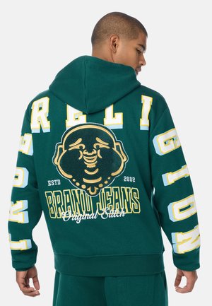 Man wearing a dark green hoodie with large yellow and white lettering, a smiling Buddha graphic, and "Brand Jeans" text on the back.