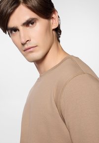 Beżowa bluza dresowa z okrągłym dekoltem, z widocznymi detalami szwów i miękką teksturą. Model przyjmuje neutralny wyraz twarzy.