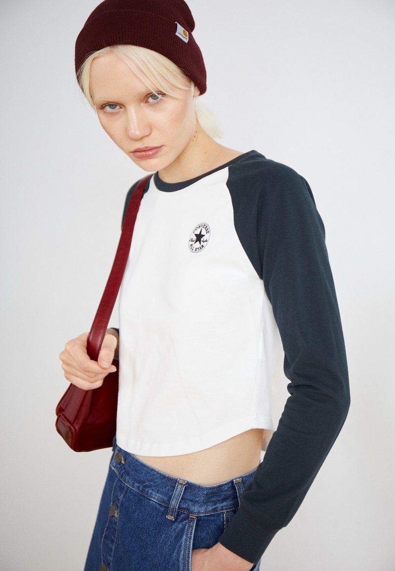 Top corto con corpo bianco e maniche lunghe verde scuro, con un patch del logo. Abbinato a una gonna di jeans e una borsa a tracolla rossa.