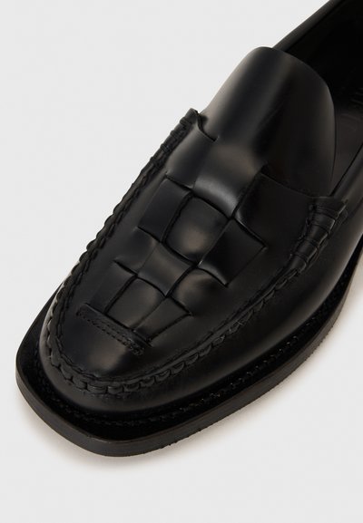 Mocassin en cuir noir avec détails tressés, texture lisse, bout rond, accents cousus et semelle noire à profil bas.