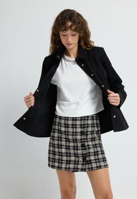 Black jacket with gold buttons and beaded accents, layered over a white top, paired with a black and white checked skirt.