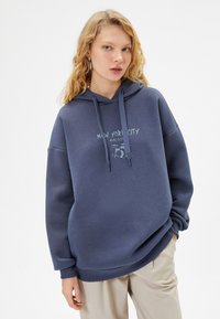 Felpa con cappuccio blu dal fit morbido, con la scritta "New York City" davanti e una grafica di un tamburo, realizzata in tessuto morbido.