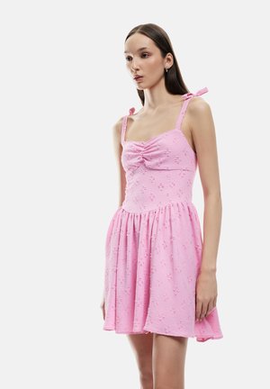 Roze jurk met textuurstof, een aansluitend lijfje en een vloeiende rok. Heeft dunne bandjes met strikjes en geplooide details op de borst.