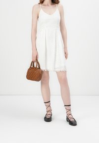 Weißes Spitzenkleid mit schwingendem Rock, Spaghetti-Trägern und gemusterter Textur. Braune gesteppte Handtasche und schwarze flache Sandalen mit Riemchen.