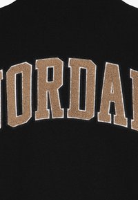 Czarna tkanina z dużymi, brązowymi, teksturowanymi literami piszącymi "JORDAN". Litery są obrysowane na biało, co tworzy kontrast z ciemnym tłem.