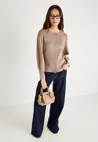 Μπεζ πλεκτό κα cardigan με στρογγυλή λαιμόκοψη και κουμπιά από πέρλες, σε συνδυασμό με φαρδιά παντελόνια από σκούρο τζιν και μια μικρή μπεζ τσάντα.