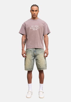 Mann mit geflochtenem Haar trägt ein verblasstes mauvefarbenes übergroßes T-Shirt, helle Jeansshorts, weiße Socken und weiße Turnschuhe und steht vor einem einfarbigen Hintergrund.