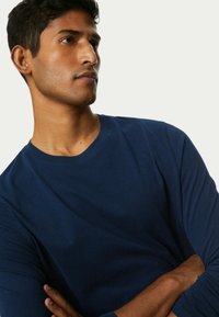 marineblaue langärmelige T-Shirt aus glattem Stoff, mit rundem Halsausschnitt und enganliegenden Ärmeln. Auf einem Modell mit verschränkten Armen gezeigt.
