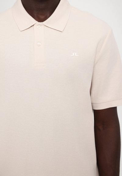 Lys beige polo-skjorte laget av strukturert stoff. Har en krage-nakke, knappelukking og en liten hvit logo på brystet.