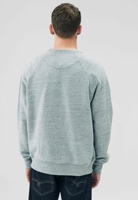 Grå sweatshirt med en struktureret strik, raglanærmer, ribbet halsudskæring og manchetter. Vist fra ryggen, parret med mørke jeans.