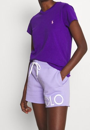 Person trägt ein lila kurzärmeliges Shirt und helllila Shorts mit weißem Kordelzug und dem Text "POLO" am linken Bein.