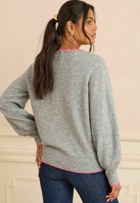 Grauer Pullover aus weichem Material, mit gepufften Ärmeln und einem rosa gewellten Saum am Halsausschnitt und Saum. Glatte Textur.