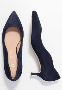 Marinblå mocka spetsiga pumps med låg klack och utskurna sidor, med en beige innersula med logotyp. Slät yta och åtsittande design.
