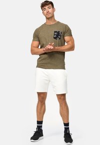 Olivengrøn t-shirt med en sort, mønstret lomme, parret med hvide shorts og sorte sneakers. Atletisk pasform og afslappet design.