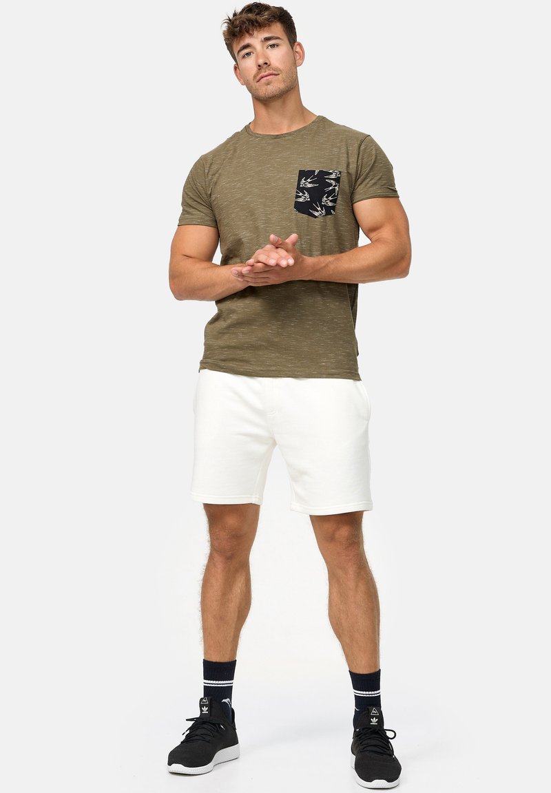 Olivengrøn t-shirt med en sort, mønstret lomme, parret med hvide shorts og sorte sneakers. Atletisk pasform og afslappet design.